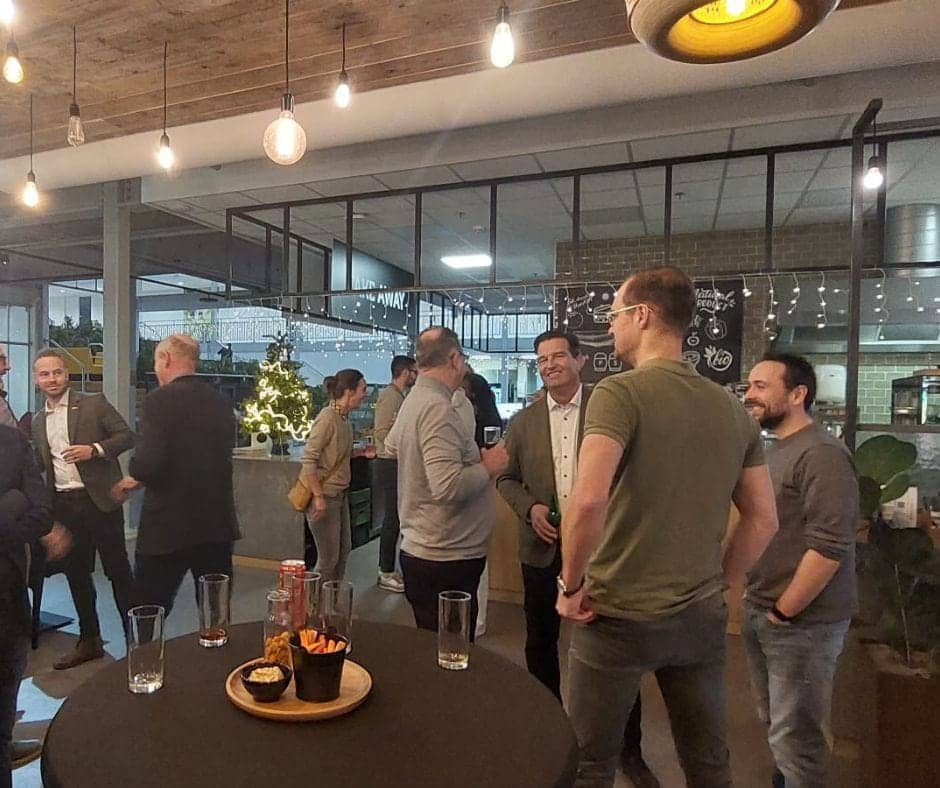 People stand around tables in an informal networking setting. They are engaged in friendly discussion.